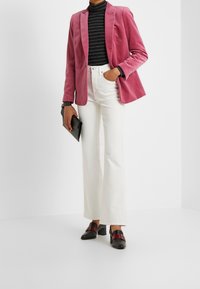 Veste en velours rose, col roulé noir à rayures, pantalons blancs évasés, chaussures à talons blocs noir et rouge, tenant une pochette noire.