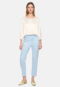 Woman wearing a loose white long-sleeve blouse, light blue fitted pants, and pale yellow pointed slingback flats, standing on a plain background.