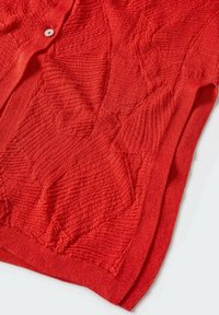 Cardigan rouge en maille avec des motifs géométriques texturés et un seul bouton blanc visible près du col sur un fond clair.