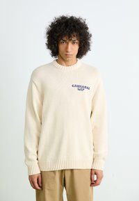 Pull en tricot couleur crème avec un col et des poignets côtelés, ornés d'un logo brodé bleu sur la poitrine gauche. Léger et texturé.