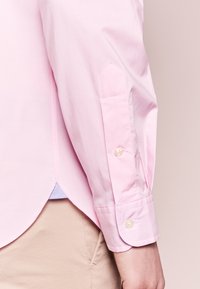 Ljust rosa skjorta i en mjuk textur, med spetsiga manschetter och en knappdetalj. Krökt nederkant synlig, lager över beige tyg.