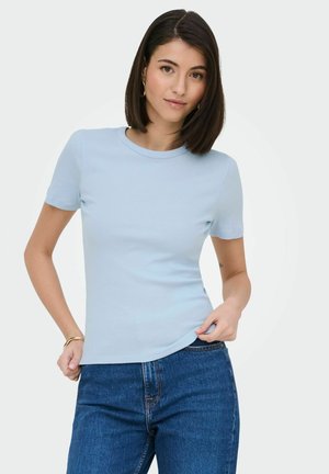 Donna con capelli lisci e scuri che indossa una maglia a maniche corte azzurro chiaro e jeans blu, che sistema l'orlo della maglia con la mano.