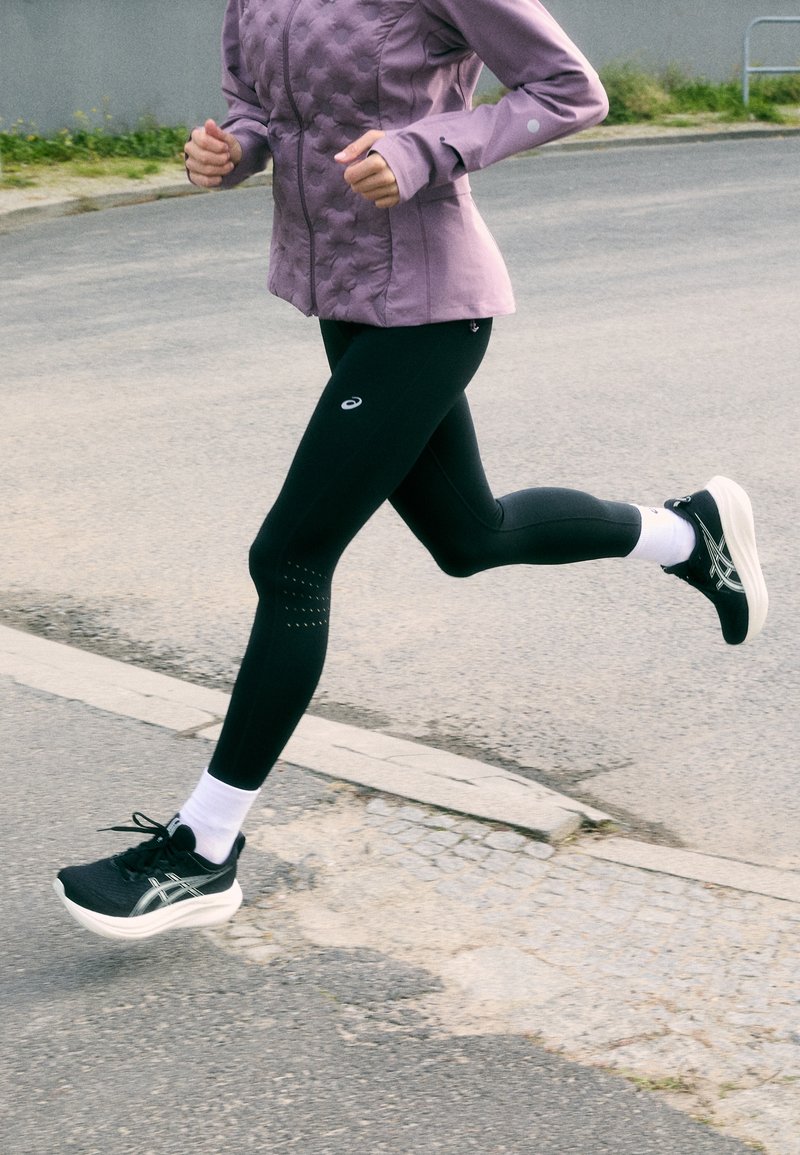 Mustad atletikud legginsid perforatsioonide detailidega, koos heleda lillaka pehme jope ja mustade jooksujalatsitega.