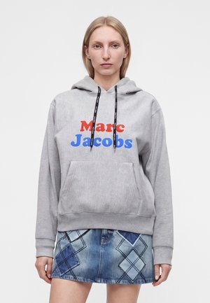 Femme portant un sweat à capuche gris Marc Jacobs avec texte rouge et bleu et une jupe en jean bleue ornée de motifs losanges cousus.