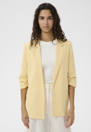 Femme aux cheveux ondulés et foncés, vêtue d'un blazer jaune pâle sur une chemise blanche et un pantalon blanc, regardant vers l'avant sur un fond uni.