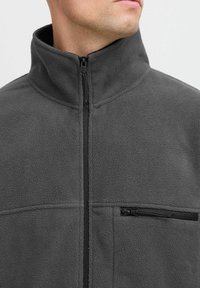 Veste en molleton gris avec un col haut et une fermeture éclair sur toute la longueur. Elle possède une poche poitrine zippée et un tissu doux et texturé.