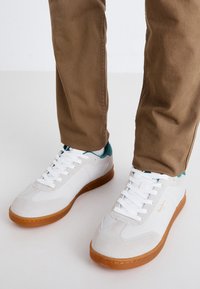 Zapatillas blancas con suelas de goma, detalles en ante y acentos en color teal en el talón, combinadas con pantalones beige. Diseño sencillo con cordones.