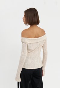 Beige off-shoulder topp med ribbet tekstur, lange tettsittende ermer, og en lett buet søm, kombinert med løse svarte bukser.