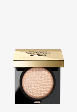 Compactpoeder in een zwarte verpakking met een goudkleurig reflecterend deksel. Het poeder binnenin heeft een zachte perzikachtige tint en een getextureerd oppervlak.