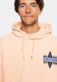 Sudadera con capucha color durazno con cordones, que presenta un logo geométrico de Quiksilver en azul y negro en el pecho. Tejido suave y texturizado.