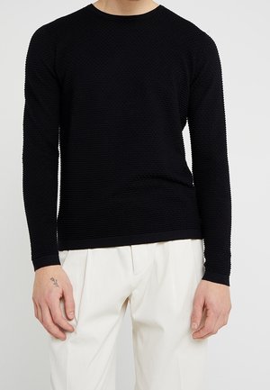 Schwarzer, langärmeliger strukturierter Strickpullover mit Rundhalskragen und gerippten Bündchen, kombiniert mit cremefarbenen, plissierten Hosen.