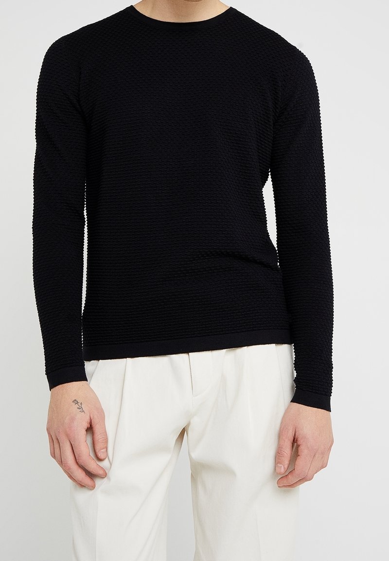 Maglione lungo nero a maniche lunghe in maglia strutturata con scollatura rotonda e polsini a costine, abbinato a pantaloni plissettati color crema.
