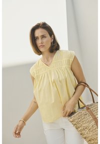 Blouse jaune sans manches avec un décolleté en V et une texture froncée, associée à un pantalon blanc et un sac en osier avec des poignées en cuir marron.