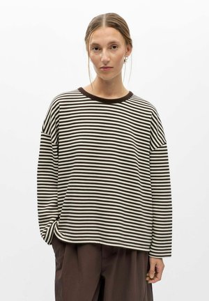 Mulher com cabelo apanhado, vestindo uma camisa de manga comprida às riscas pretas e brancas e calças castanhas largas, de pé contra um fundo branco simples.