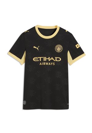 Kvinde iført en sort og gul Manchester City fodboldtrøje med et langt blåt denimskørt, der holder en sort håndtaske mod en grå baggrund.