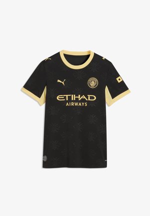 Mujer con camiseta de fútbol del Manchester City negra y amarilla, con una falda larga de mezclilla azul, sosteniendo un bolso negro frente a un fondo gris.