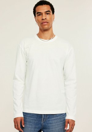 Mann mit kurzen lockigen Haaren, der ein weißes Langarmshirt, eine silberne Kettenhalskette, Creolen und blaue Jeans vor einem schlichten Hintergrund trägt.