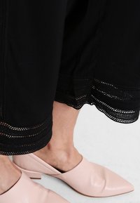 Svarta byxor med dekorativa snören vid fållen, tillsammans med ljusrosa läderklackar med spetsig tå och öppen baksida.