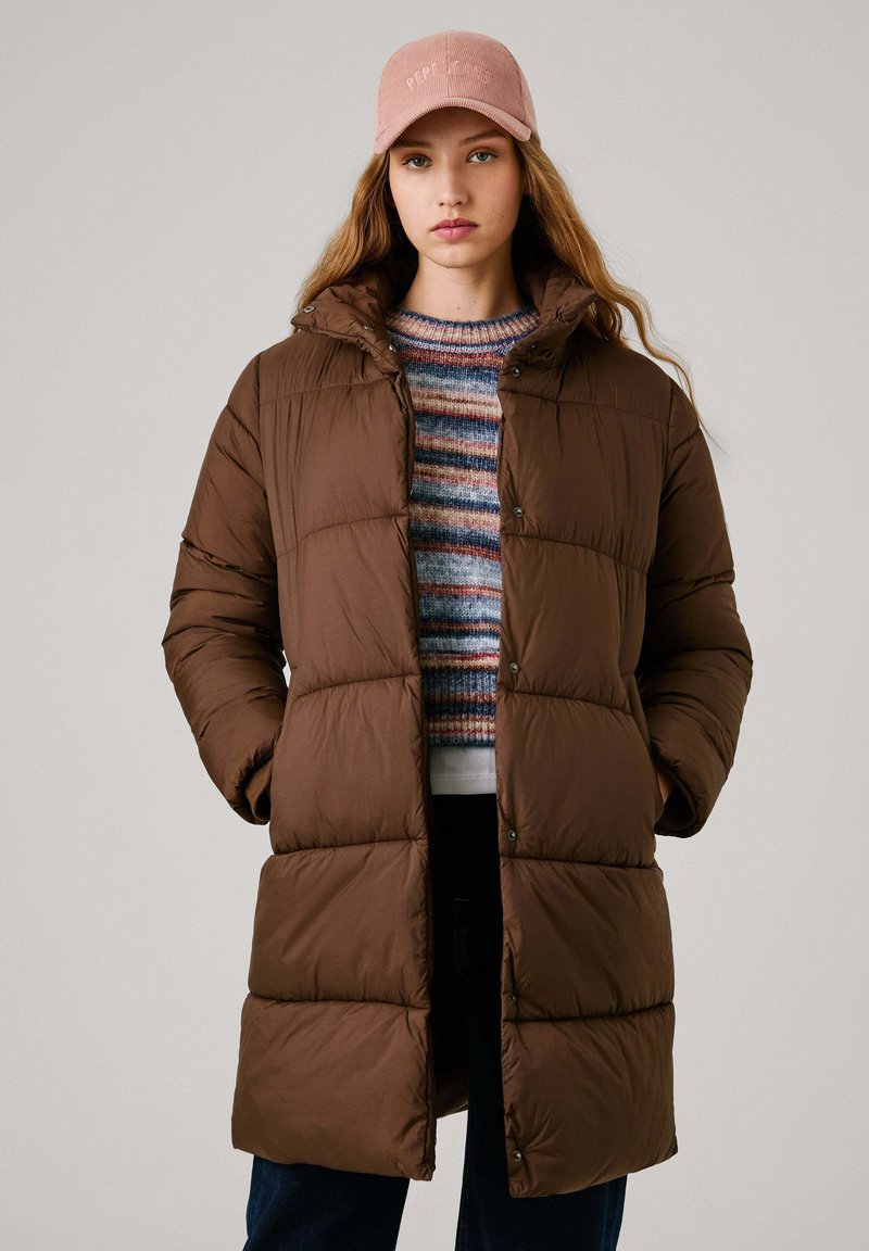 Brązowa kurtka puchowa z kapturem, charakteryzująca się pikowanym wzorem i zapięciem na napy. Nosi ją osoba w kolorowym swetrze w paski i ciemnych dżinsach.