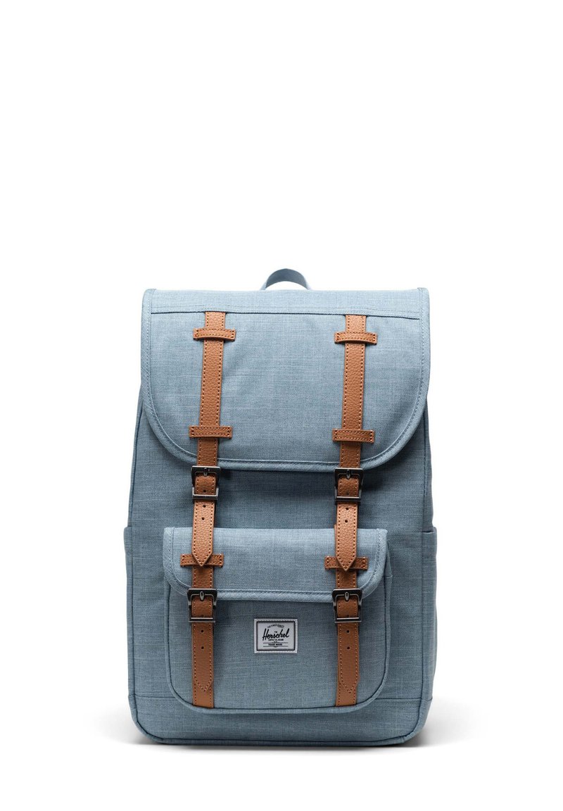 Blå tygryggsäck med bruna läderremmar, två framfickor med spännen, övre flik och en central logotypapplikation. Slät textur, strukturerad design.