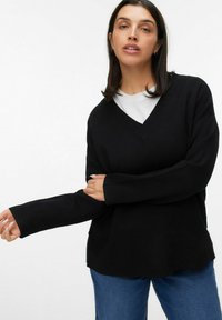Czarny sweter z dekoltem w serek, z żebrowanym wykończeniem, długimi rękawami i luźnym fasonem, noszony na białej koszulce; zestawiony z niebieskimi dżinsami, prezentowany na jasnym tle.