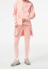 Ljusrosa zip-up hoodie med framfickor och ribbade ärmslut, tillsammans med matchande sweatpants med sidoklipper. Bärs med grå sneakers.