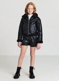 Schwarze Kunstlederjacke mit einem Schafslederkragen, cropped Design, mit Reißverschluss versehenen Taschen, kombiniert mit schwarzen Lederhotpants und Schnürstiefeln.