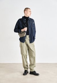 Farmering gombos ing, fekete garbó, olajzöld cargo nadrág, fekete cipő és egy kis barna keresztpántos táska. Semleges háttér.