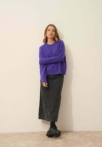 Lila gestrickter Pullover mit gerippten Bündchen, kombiniert mit einem langen schwarzen Jeansrock und schwarzen klobigen Schuhen. Glatte Textur, lässiges Design.