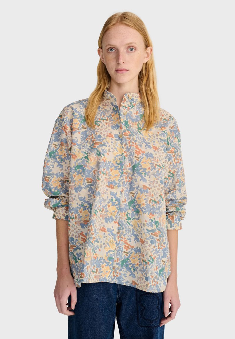 Jeune femme aux cheveux blonds raides porte une chemise ample à motifs multicolores boutonnée et un pantalon bleu foncé sur un fond uni.