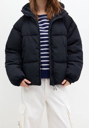 Femme portant une doudoune bleu marine par-dessus un pull à rayures bleu marine et blanc et un pantalon cargo blanc, debout avec les mains dans les poches.