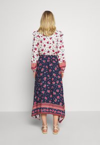 Blouse fleurie avec épaules volantées, associée à une jupe bleu marine ornée d'un motif floral rouge et d'un ourlet décoratif. Sandales marron clair complètent la tenue.