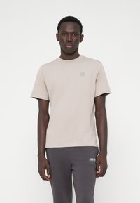 Ανοιχτό μπεζ βαμβακερό t-shirt με στρογγυλή λαιμόκοψη και κοντά μανίκια. Διαθέτει ένα μικρό λογότυπο σε πιο σκουρόχρωμη απόχρωση στην περιοχή του στήθους.