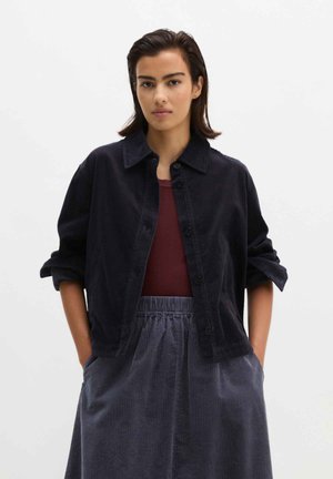 Femme portant une veste en velours côtelé foncé sur un haut bordeaux et un pantalon large en velours côtelé assorti, les mains dans les poches.