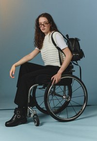 Fehér kötött ujjatlan mellény, fehér pólóval, fekete, bő szárú nadrággal és fekete, csatos bokacsizmával párosítva. Egy fekete hátizsák pihen a kerekesszéken.