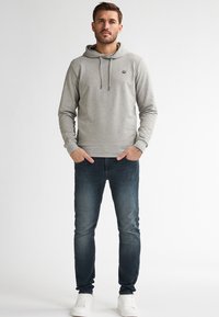 Hombre de pie con las manos en los bolsillos, vistiendo un hoodie gris claro, jeans oscuros deslavados y zapatillas blancas, contra un fondo blanco.