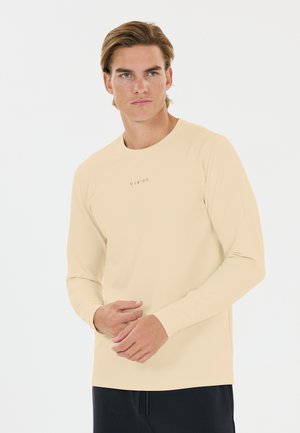 Ung mand med lyst brunt hår iført beige langærmet skjorte med teksten "VIRTUS" og sorte bukser mod en hvid baggrund.