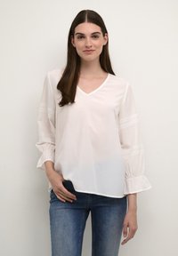 Camicia bianca con scollo a V, maniche lunghe con polsini a volant e dettagli in pizzo sulle maniche. Abbinata a jeans in denim blu.