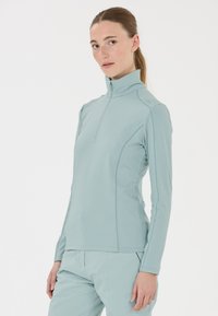 Vaaleansininen pitkähihanen toppi, jossa on korkea kaulus ja osittainen vetoketju. Takki on valmistettu sileästä ja joustavasta kankaasta, ja siinä on hienovaraisia sauman yksityiskohtia.