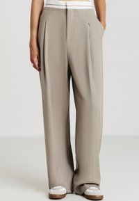 Personne portant un pantalon beige taille haute évasé avec des plis, des détails de ceinture blanche et des baskets blanches, se tenant les mains dans les poches.