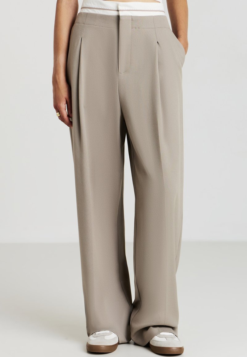 Personne portant un pantalon beige taille haute évasé avec des plis, des détails de ceinture blanche et des baskets blanches, se tenant les mains dans les poches.