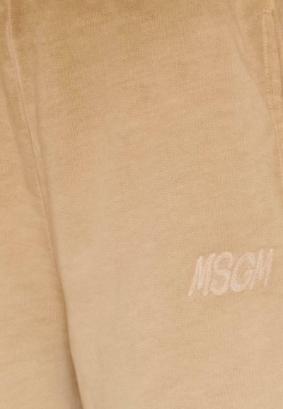 Beige sweatpants gemaakt van zachte stof met een geborduurd logo, zichtbare stiksels en een ontspannen pasvorm. Geen zichtbare zakken.