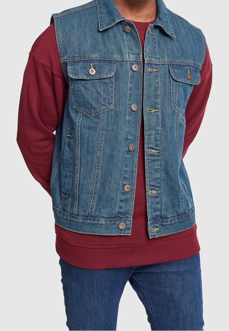 Jeansväst i blått, med två bröstfickor, metallknappar och fransad nederkant. Lagd över en vinröd sweatshirt, med blå jeans synliga nedanför.