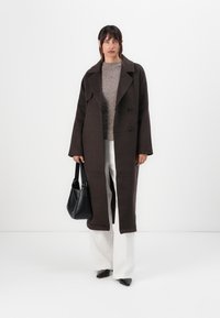 Long brown coat with a double-breasted design, wide lapels, and a structured fit. Worn over a light gray sweater and white trousers. Black handbag.