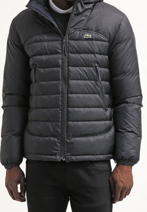 Zwarte donzen jas met ritssluiting aan de voorkant en met rits afgesloten zijzakken, gedragen door een persoon met de handen ontspannen langs het lichaam.