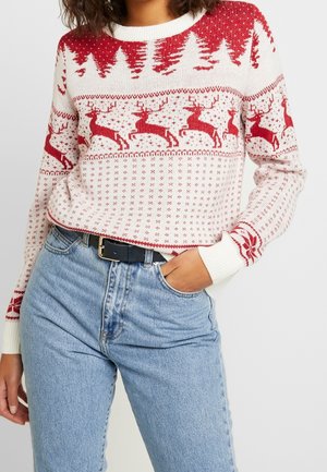 Pull en tricot blanc avec des motifs de sapins de Noël rouges et de rennes, porté avec un jean bleu clair et une ceinture noire.