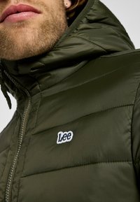 Grön vadderad jacka med hög krage, dragkedjestängning och broderad "Lee"-logga på bröstet. Slät, glansig textur och sydde mönster.