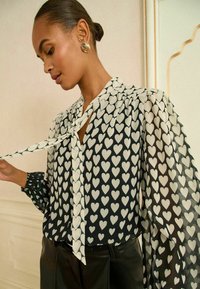 Blusa nera decorata con motivi a cuore bianchi, con chiusura al collo annodata e maniche trasparenti e texturizzate con polsini arricciati.