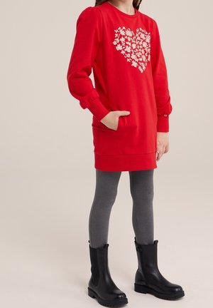 Niña con vestido sudadera rojo con un corazón floral blanco, leggings grises y botas negras hasta el tobillo, de pie con una mano en el bolsillo.