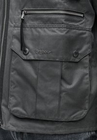 Τσέπη σκούρου γκρι μπουφάν με φερμουάρ και καπάκι με κουμπιά πατιλέτα, με κεντημένο λογότυπο "Barbour" στο ύφασμα.